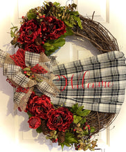 Load image into Gallery viewer, This is our grapevine welcome wreath it is approximately 24 inches in circumference and it has beautiful red flowers with greenery and berries on it. It has a homemade sash that says welcome on it in a checkered black and white with red lettering on it and it’s perfect for any door or wall. This beautifully handmade bow on this wreath makes it say “Welcome” and it is gorgeous!￼