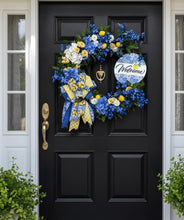 Load image into Gallery viewer, Our stunning handcrafted wreath is designed on a natural grapevine base and beautifully adorned with an elegant arrangement of premium silk florals and vibrant seasonal accents. Featuring gorgeous blue hydrangeas, delicate blue delphinium blooms, soft ivory and buttery yellow ranunculus, and lush greenery, this design captures the perfect balance of sophistication and cheerful summer charm.
Bright yellow lemons are throughout the wreath, adding a fresh pop of color and a timeless Mediterranean touch.