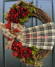 Load image into Gallery viewer, This is our grapevine welcome wreath it is approximately 24 inches in circumference and it has beautiful red flowers with greenery and berries on it. It has a homemade sash that says welcome on it in a checkered black and white with red lettering on it and it’s perfect for any door or wall. This beautifully handmade bow on this wreath makes it say “Welcome” and it is gorgeous!￼